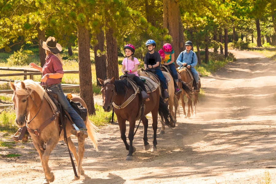 Family horseback riding in Montana on a dirt road, with a wrangler leading the way