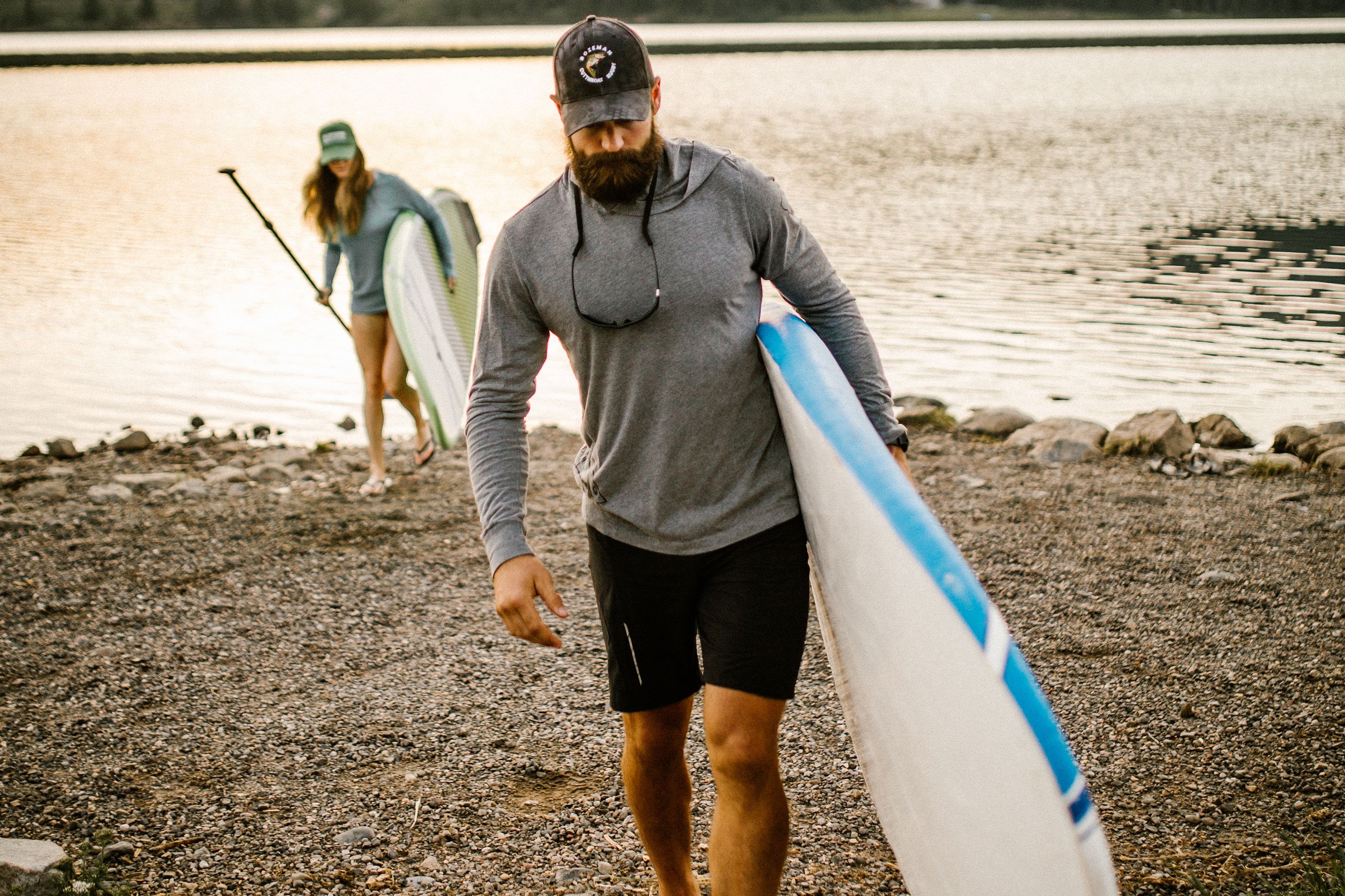 Man and woman carrying standup paddle boards from lake. 